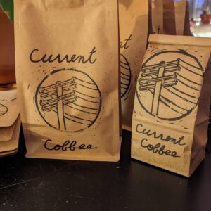 One 1/2lb bag of coffee next to a 1/4lb bag of coffee on a black table inside a house with a green monstera in the back and a window looking outside.