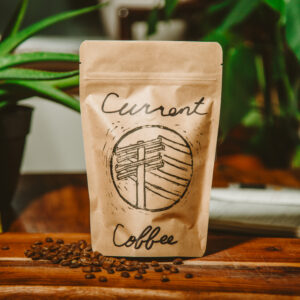 A bag of roasted coffee sitting on a wooden board with roasted coffee beans sprinkled around and a aloe plant in the background.